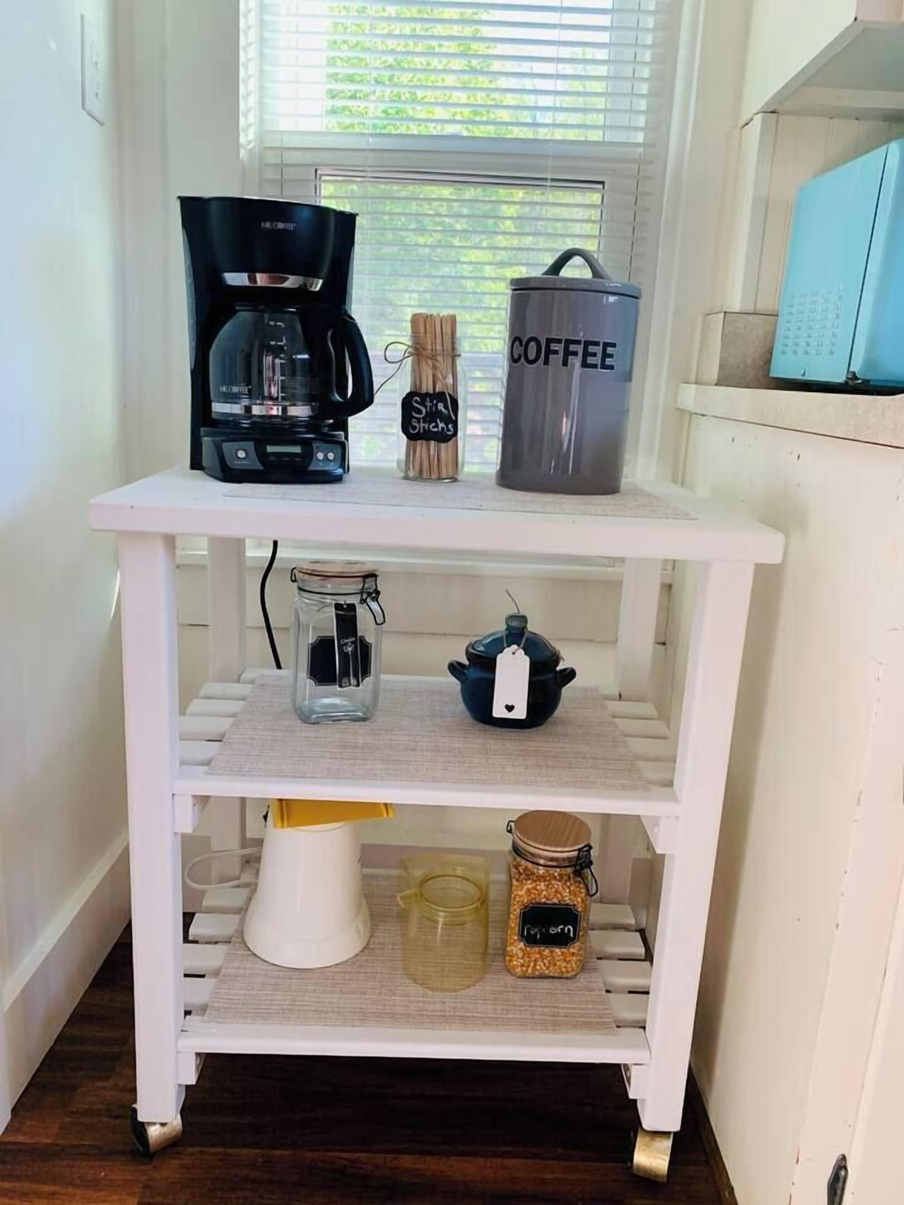 A white kitchen cart with a coffee maker, a jar of spices, and a jar of popcorn.