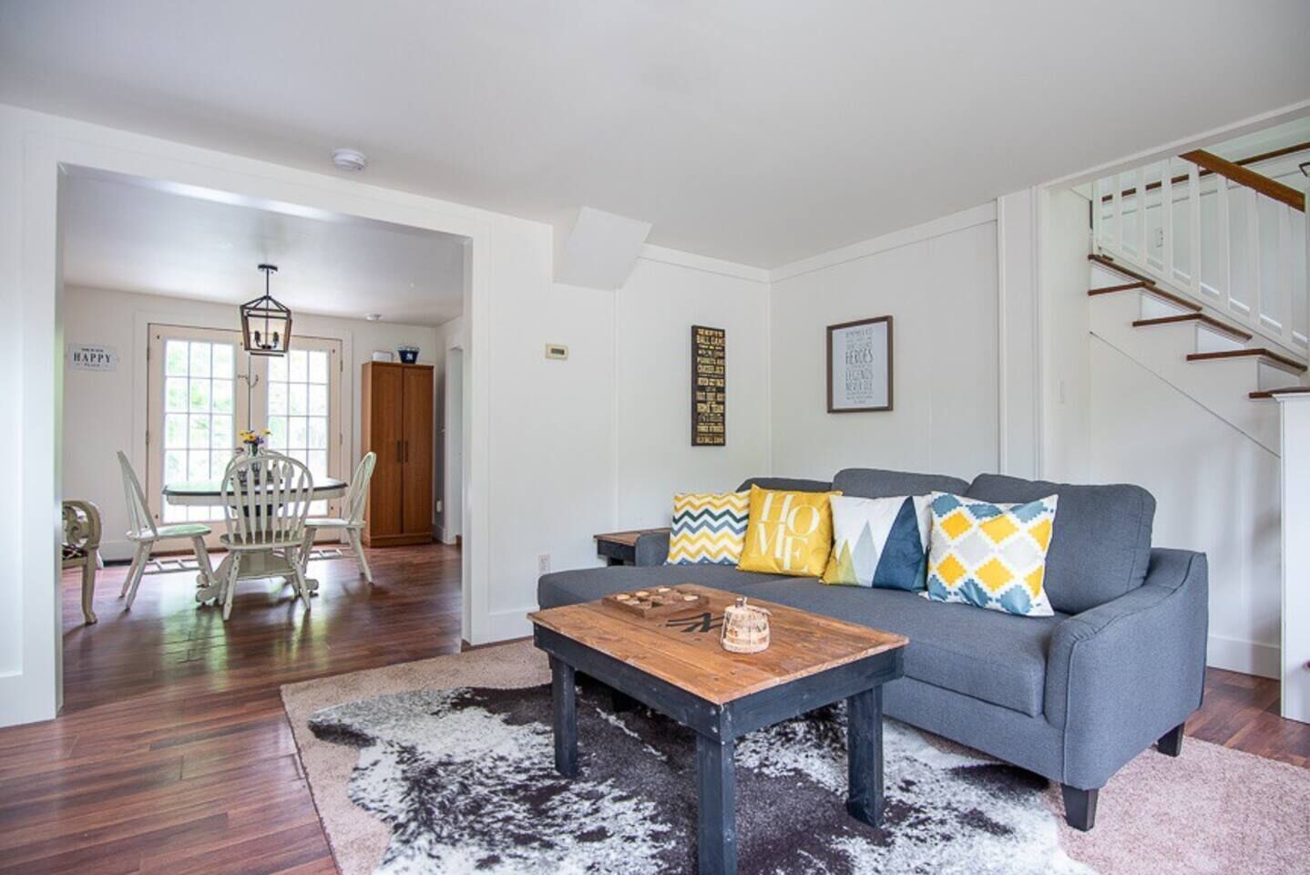 Ein Wohnzimmer mit grauem Sofa, einem Holztisch und gemusterten Kissen. Rechts ist eine Treppe zu sehen, und im Hintergrund befindet sich ein Essbereich mit weißen Stühlen und einem Holzschrank.