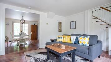 Ein Wohnzimmer mit grauem Sofa, einem Holztisch und gemusterten Kissen. Rechts ist eine Treppe zu sehen, und im Hintergrund befindet sich ein Essbereich mit weißen Stühlen und einem Holzschrank.