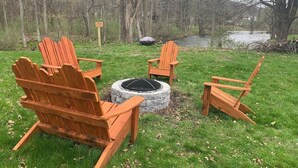 Een ronde vuurkuil van steen met vier houten Adirondack-stoelen eromheen, gelegen in een gazon met bomen en een waterpartij op de achtergrond.