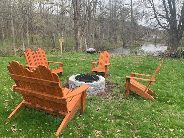 Un pozo de fuego circular de piedra con cuatro sillas Adirondack de madera a su alrededor, ubicado en un área cubierta de césped con árboles y una masa de agua al fondo.