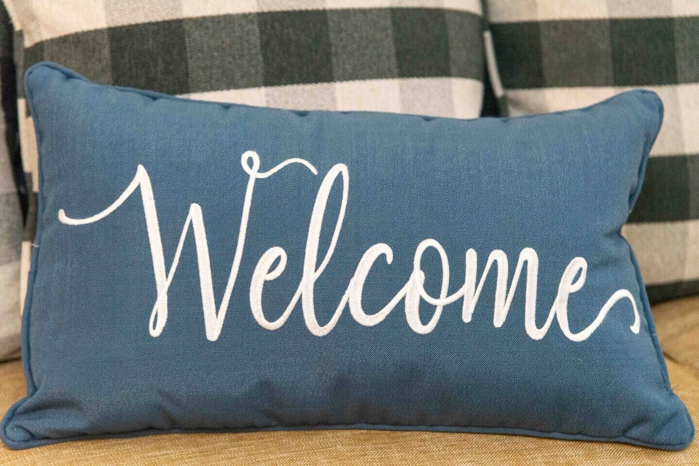 Een blauw rechthoekig kussen met de tekst 'Welcome' geborduurd in witte sierletters.