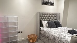 Un dormitorio con un cabecero gris con textura, una cama con ropa de cama floral, un recipiente de almacenamiento de plástico transparente, una cesta de mimbre y una obra de arte con un paisaje urbano en la pared.