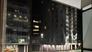 Vista nocturna desde una ventana sobre un estacionamiento con varios coches, un edificio con un letrero 'BBC' y una cafetería 'COSTA'.