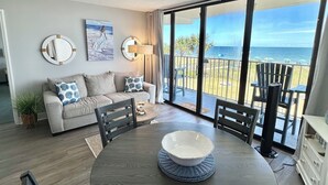A modern living room with a round dining table, a sofa, and a large window overlooking the ocean.