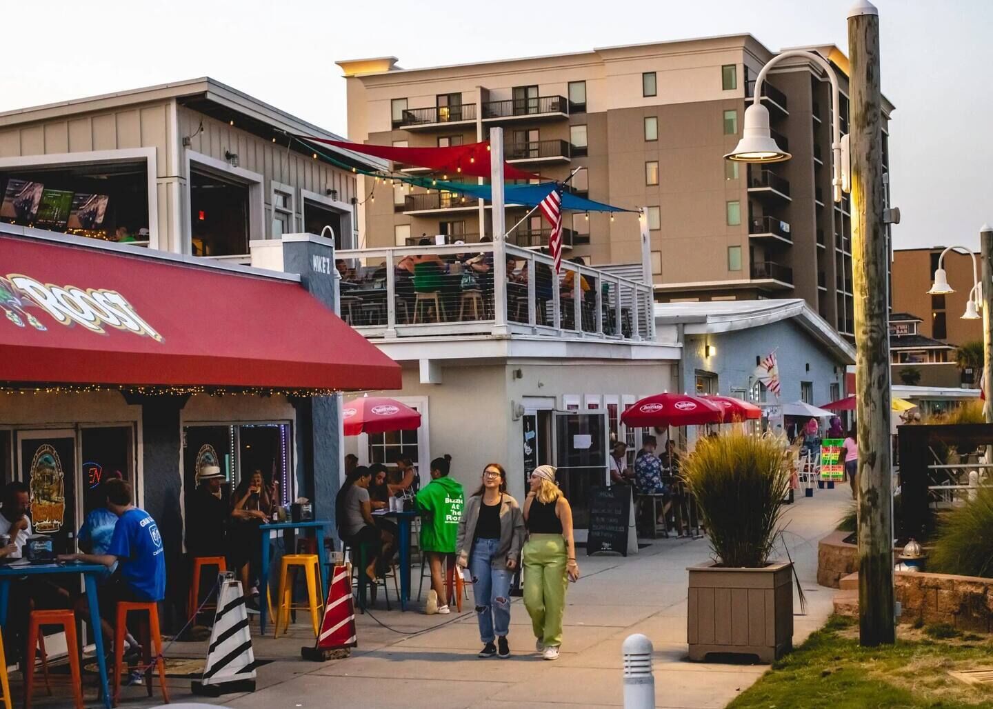 A bustling outdoor dining area with multiple tables and umbrellas, a building with balconies, and a street lined with various shops and eateries.