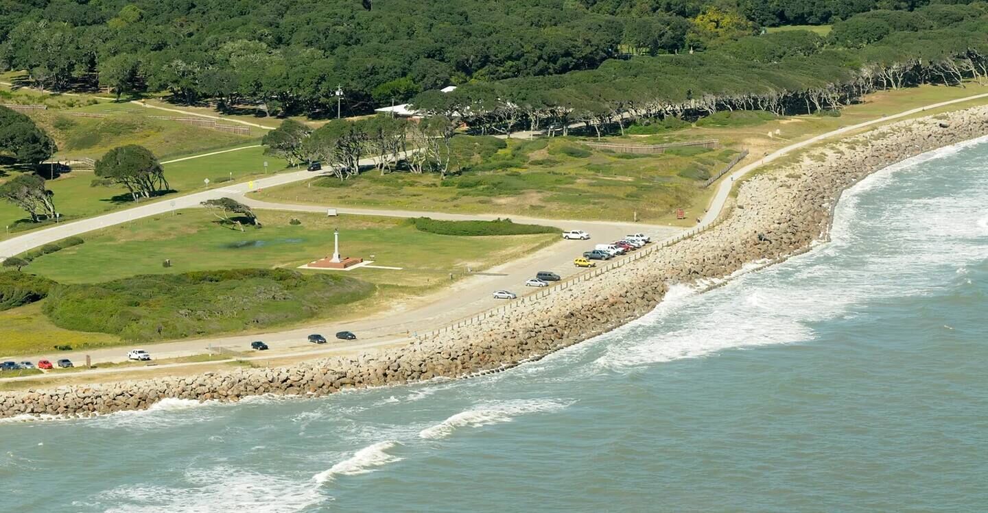 A coastal road with a rocky breakwater, a grassy area with a monument, and parked cars along the shore.