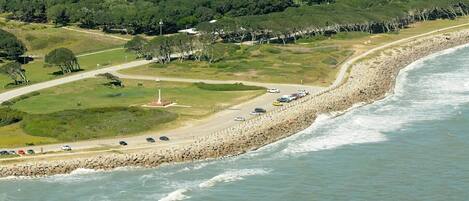 A coastal road with a rocky breakwater, a grassy area with a monument, and parked cars along the shore.