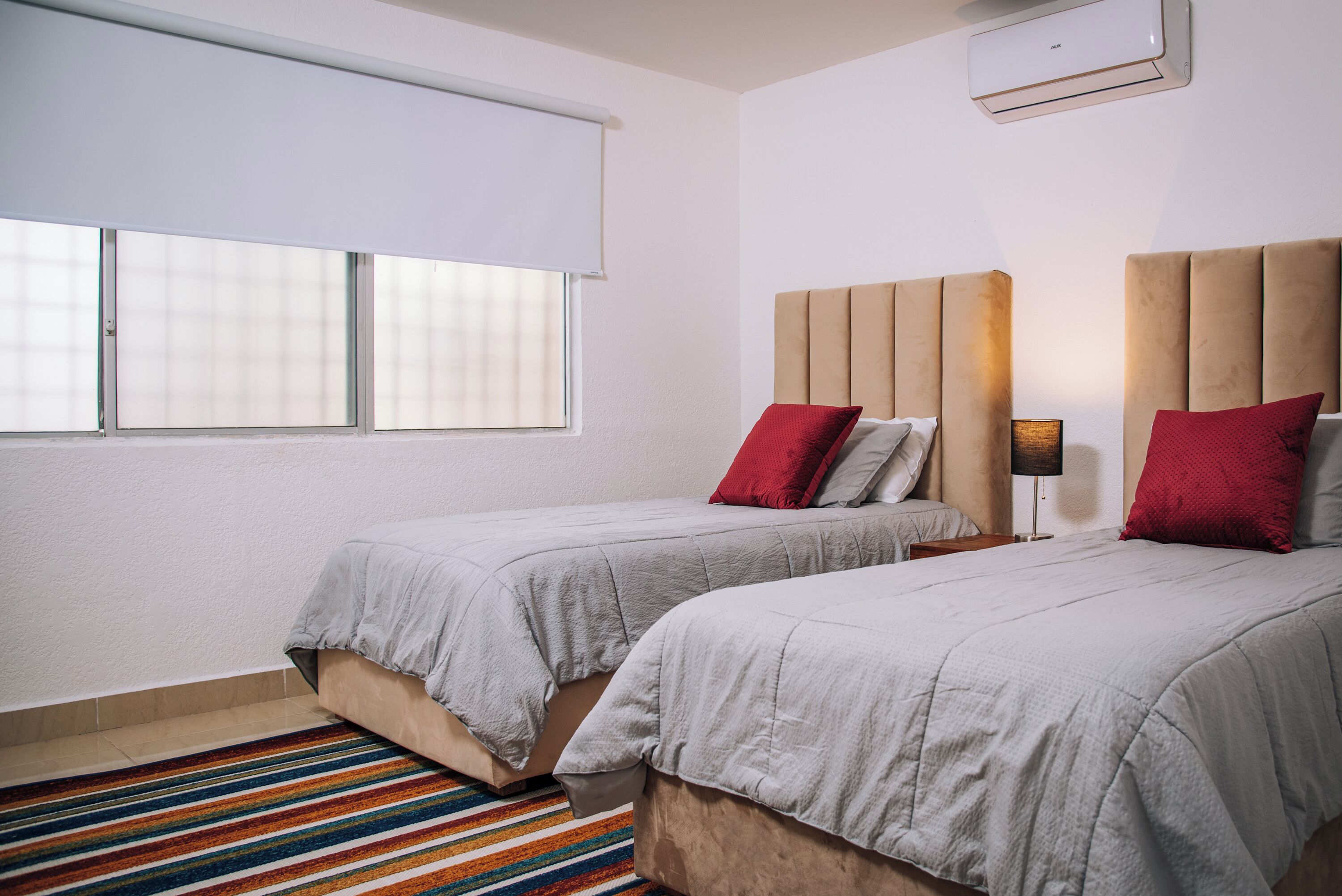 Ett hotellrum med två sängar, en randig matta, ett fönster med persienner och en väggmonterad luftkonditioneringsanläggning.