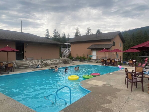 A swimming pool with a few people, surrounded by outdoor seating and umbrellas, with a building in the background. - Mara Spring Lodges - Suite 2 (Mara)