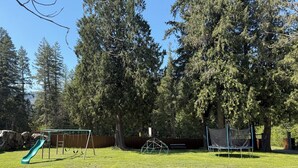 A playground with a slide and swings, surrounded by trees and a wooden fence. - Mara Spring Lodges - Suite 2 (Mara)