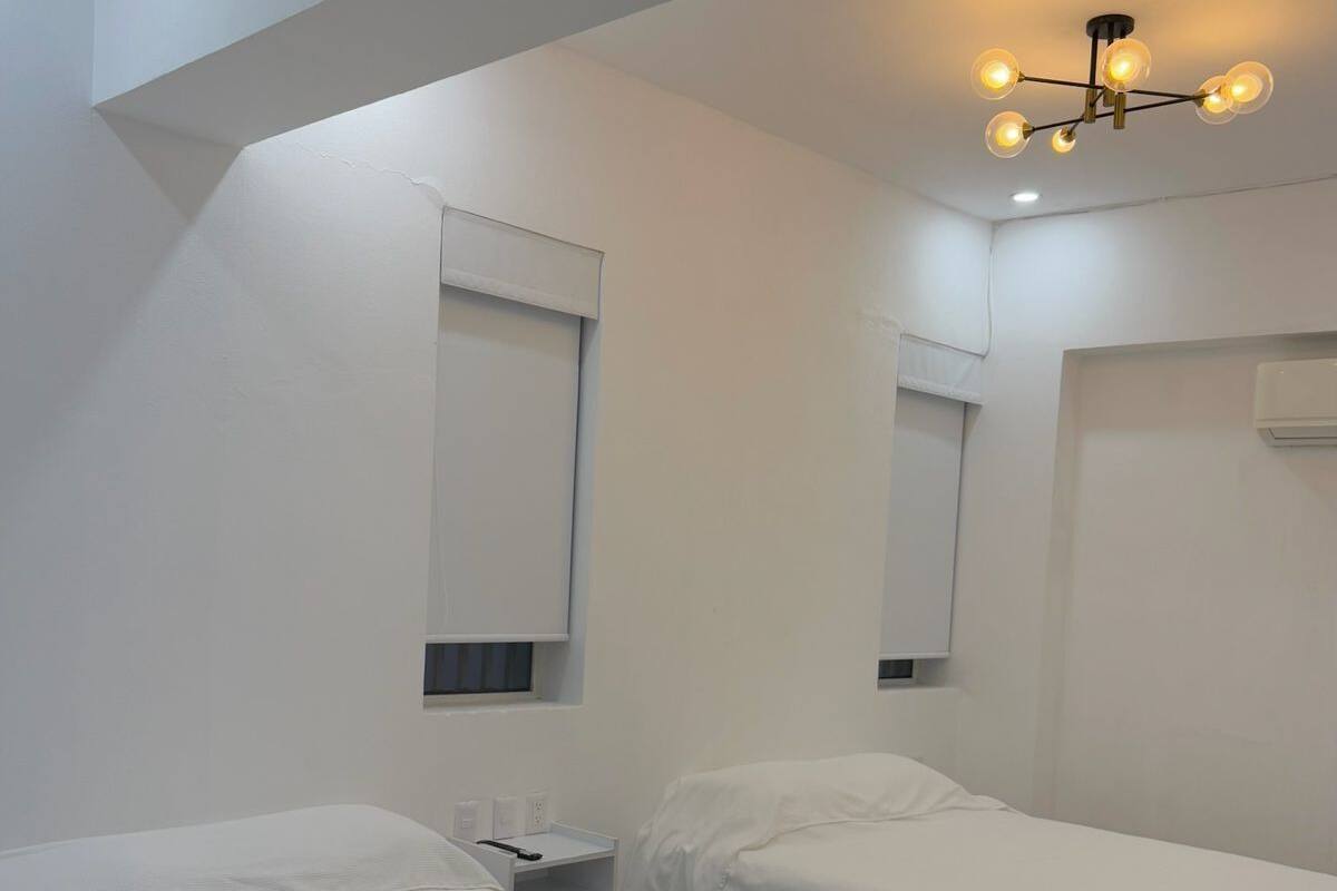 A minimalist hotel room with two single beds, a small bedside table, a wall-mounted air conditioning unit, and a modern ceiling light fixture.