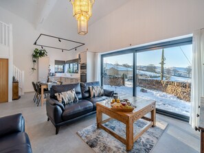 A modern living room with a black leather sofa, a wooden coffee table with snacks, and a large window overlooking a snowy landscape.