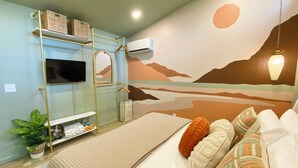 Ein Schlafzimmer mit Bett, Kissen, Fernseher, Spiegel, einer Regalreihe mit Körben, einer Pflanze und einem Wandbild.