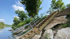 Eine hölzerne Treppe mit Metallgeländern führt zu einem Flussufer, umgeben von großen Steinen und viel Grün.