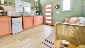 Eine Küche mit Holzausstattung, einem weißen Kühlschrank, einer Mikrowelle und einem Spülbecken. Ein Korbstuhl und ein kleiner runder Tisch mit einer Schüssel darauf sind ebenfalls vorhanden.