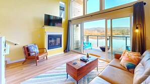 A spacious living room with a fireplace, a sofa, a coffee table, and a large sliding glass door leading to a balcony with a view of a lake. - Pond View At The Commons - Dog Friendly (Seaside)