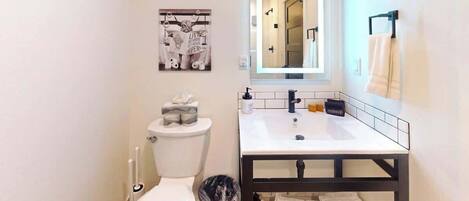 Un baño con inodoro, lavabo y espejo. Hay un perchero en la pared y un cuadro sobre el espejo.