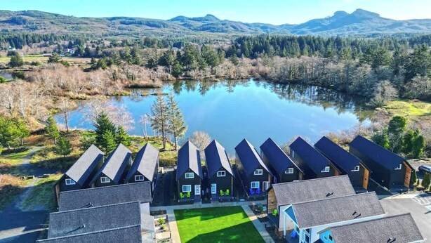 A row of modern, dark-colored cabins with pitched roofs, arranged in a line beside a calm lake, surrounded by forested hills. - Courtyard View At The Commons- 21 (Seaside)