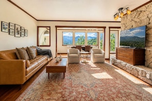 A living room with a brown leather sofa, a coffee table, two armchairs, a TV on a stone wall, and a large window with a mountain view.