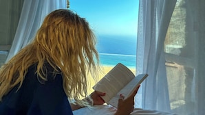 A person reading a book by a window with sheer curtains, overlooking a beach. - AGADA Folegandros (Folegandros)