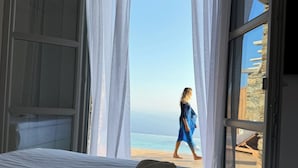 A hotel room with a bed, a hat, and sunglasses on the bed. There is a view of the beach and ocean through the window. - AGADA Folegandros (Folegandros)