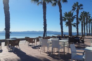 An outdoor seating area with tables and chairs, palm trees, and a view of the sea and mountains. - 3 Bed Apartment Sleeps 6 With Shared Pool (San Javier)