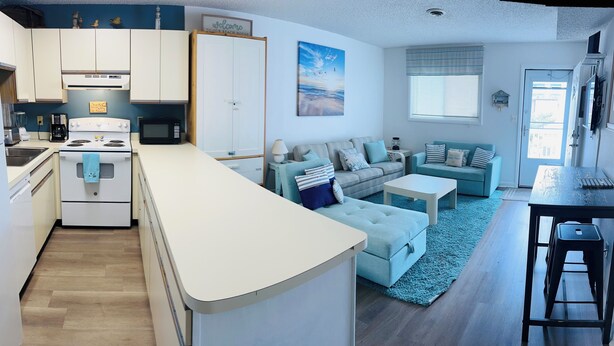 A compact kitchen with white cabinets, a stove, and a coffee maker. There is a seating area with a blue sofa and a striped couch. A painting of a beach is on the wall, and a window provides natural light. - Relisted!Ocean Blk, Bay&Ocean view, steps to beach best food & bars 2 Full Baths (Ocean City)