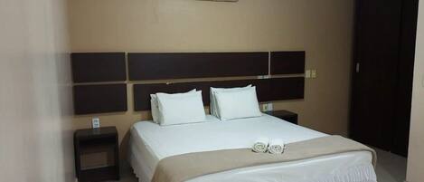 A hotel room with a bed, two bedside tables, a headboard, and an air conditioning unit.