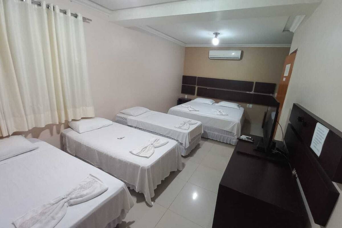 Quarto de hotel com quatro camas de solteiro, uma escrivaninha com computador e um ar condicionado na parede.