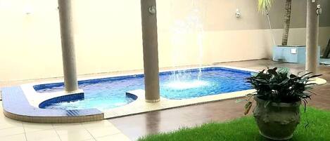 Un espacio de spa con un jacuzzi y una pequeña fuente, rodeado por un suelo de baldosas y una zona ajardinada con una planta en maceta.