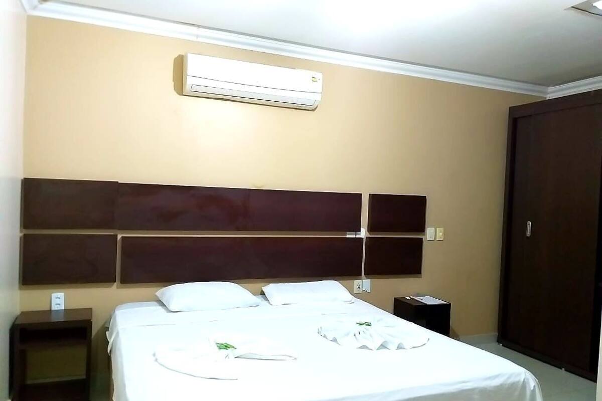 Quarto de hotel com uma cama grande, cabeceira de madeira, ar condicionado, mesa de cabeceira e guarda-roupa.