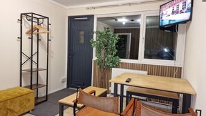 A compact living space with a wooden table and chairs, a black metal coat rack, a yellow ottoman, a potted plant, and a TV mounted on the wall.