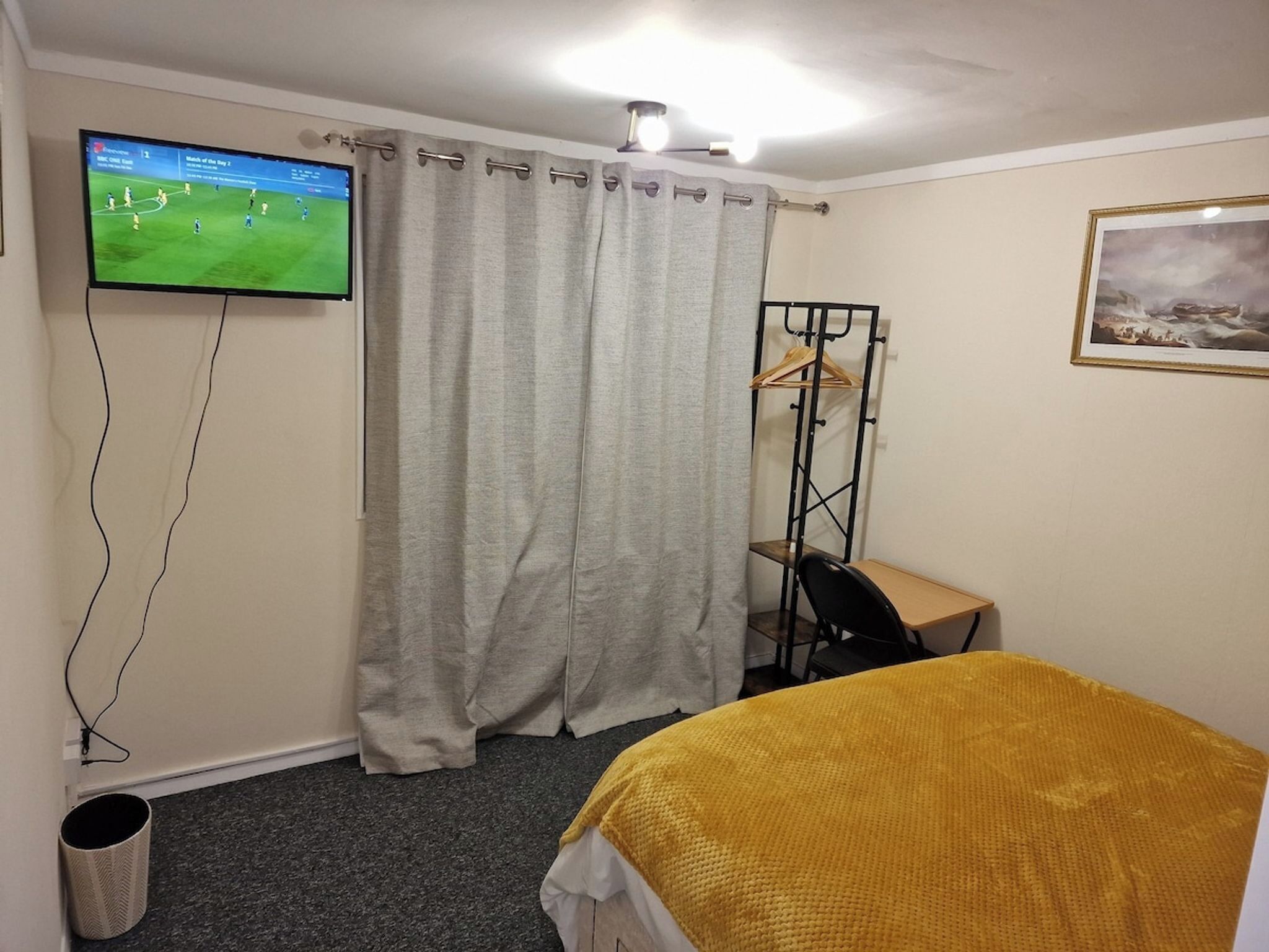 A hotel room with a bed, a desk, a chair, a television mounted on the wall, a curtain, and a framed picture on the wall.