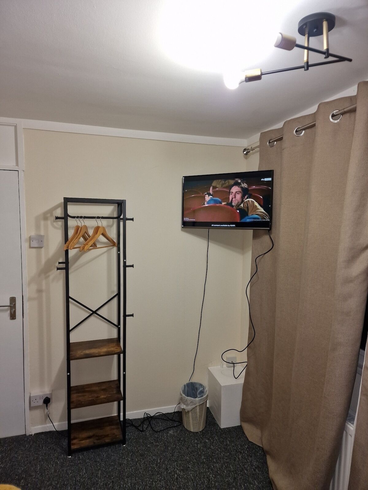 A room with a TV mounted on the wall, a wooden shelf unit, a trash bin, and a curtain.