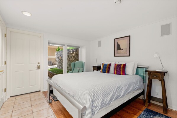 A bedroom with a bed, a chair, a bedside table, a picture on the wall, and a door leading to an outdoor area.
