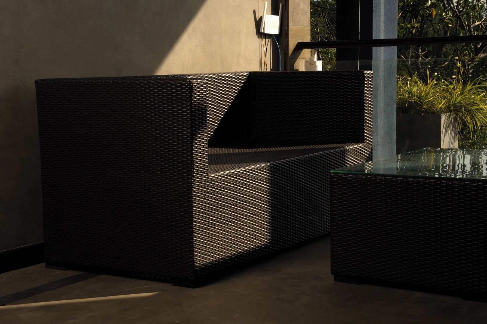 A modern outdoor seating area with a woven design, a glass table, and a view of greenery.
