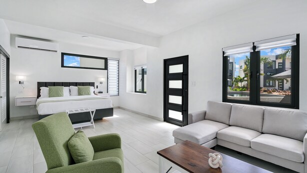 Habitación de hotel moderna con una cama, un sillón verde, un sofá gris y vistas al exterior.