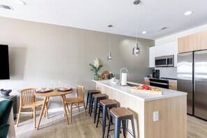 Cocina moderna con una isla central, taburetes de bar, sillas de madera, microondas y refrigerador de acero inoxidable.