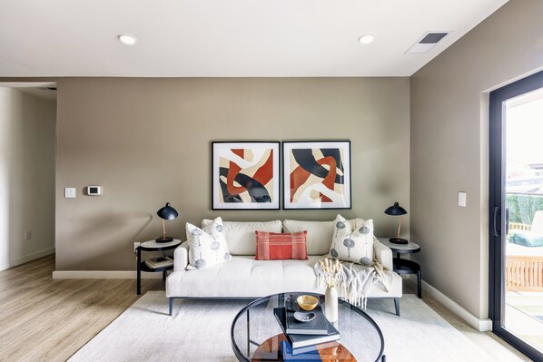 A modern living room with a white sofa, two side tables, abstract art on the wall, and a large window.