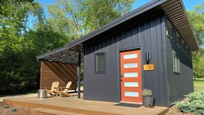 A small, modern cabin with a wooden deck, two Adirondack chairs, and a red door.