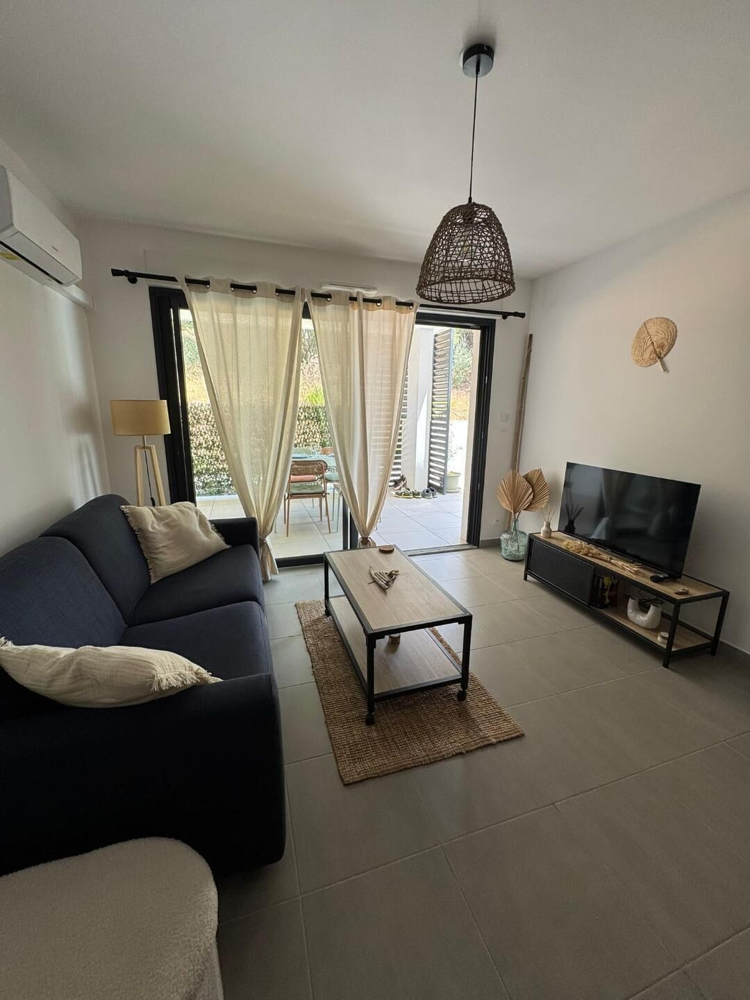 Uma sala de estar com sofá, mesa de centro, painel para TV e uma janela grande com cortinas.