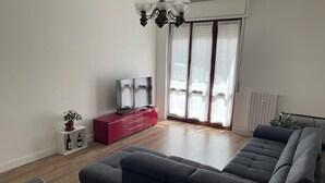 Una sala moderna con un amplio sofá seccional gris, un mueble de entretenimiento rojo con televisor de pantalla plana, una mesa de comedor con sillas y un ventanal con cortinas traslúcidas.