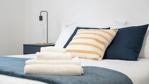 Una cama bien hecha con ropa de cama blanca y azul, una almohada a rayas y una lámpara de noche.