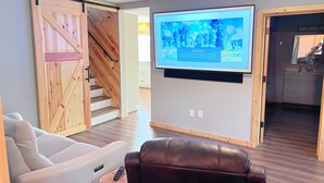 Uma sala de estar moderna com uma televisão de tela plana grande fixada na parede, uma escada de madeira, um sofá branco, uma poltrona de couro e um banquinho de madeira com um controle remoto.