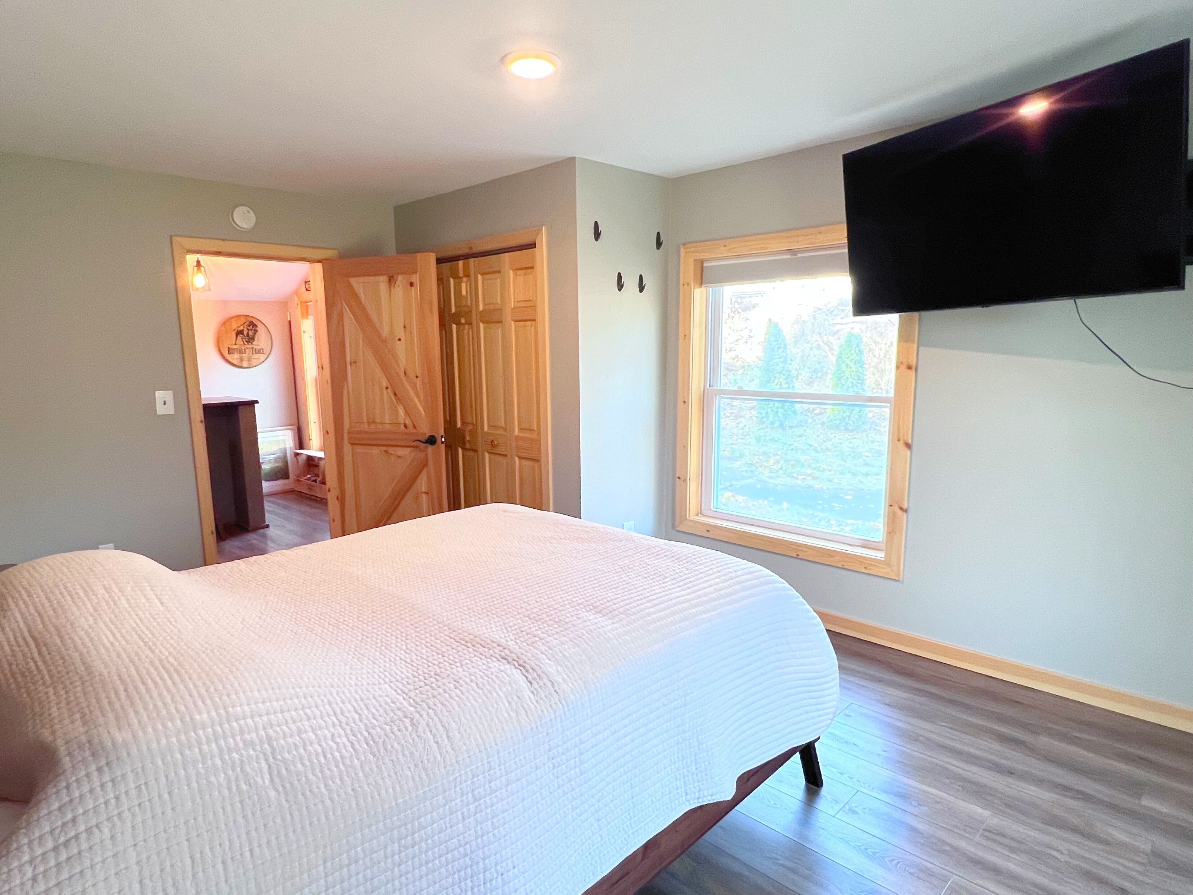 A bedroom with a large bed, a TV mounted on the wall, a window with a view of trees, and a door leading to another room.