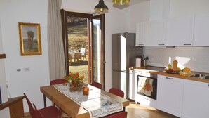 A dining area with a table set for four, a kitchen with white cabinets, and a view of a garden through the open door. - Beautiful cottage a few miles from Bellagio with stunning views of Lake Como (Civenna di Bellagio)