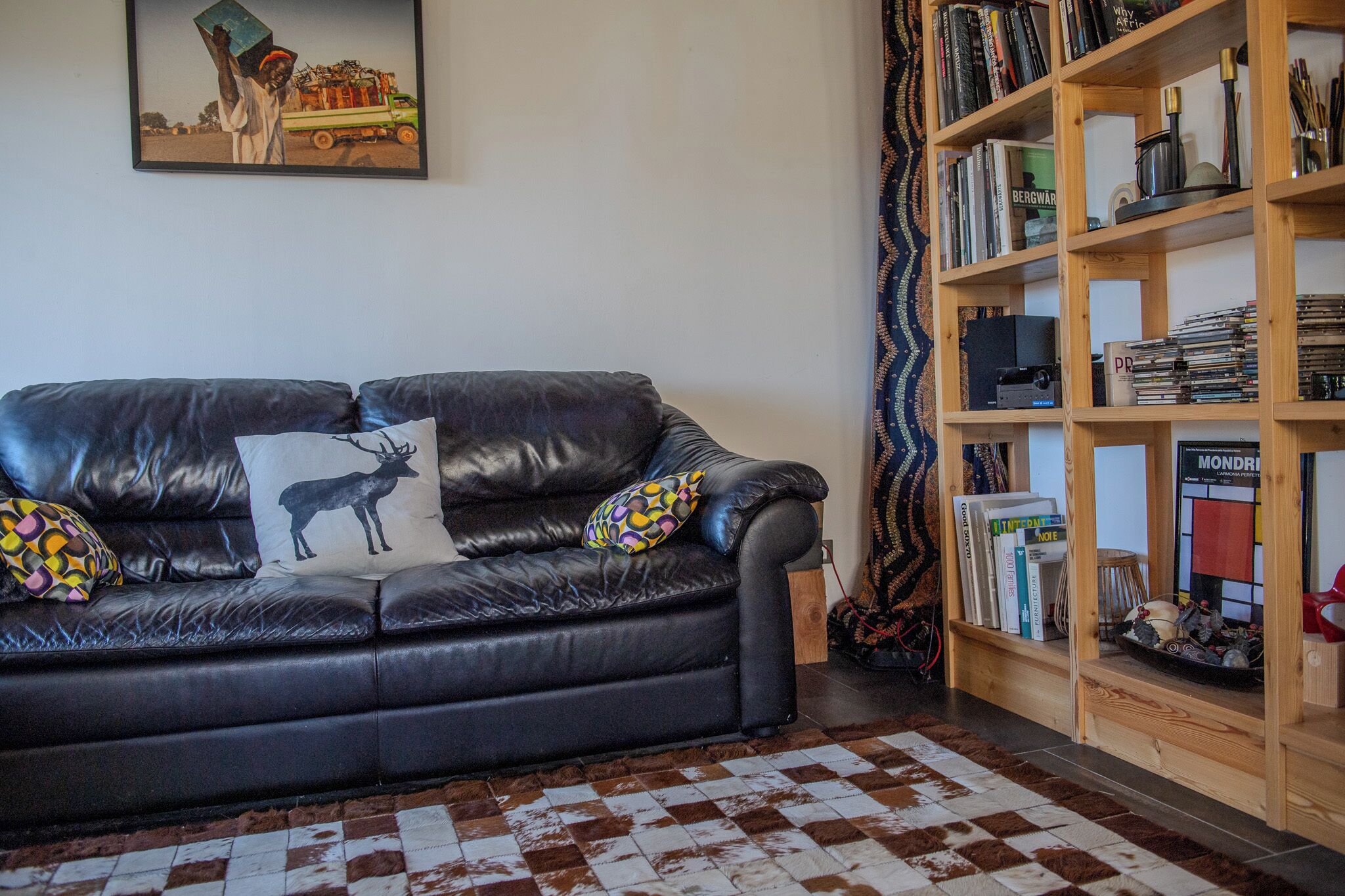 Een zwarte leren bank met decoratieve kussens, een vloerkleed met een patroon en een houten boekenkast vol boeken en decoraties.