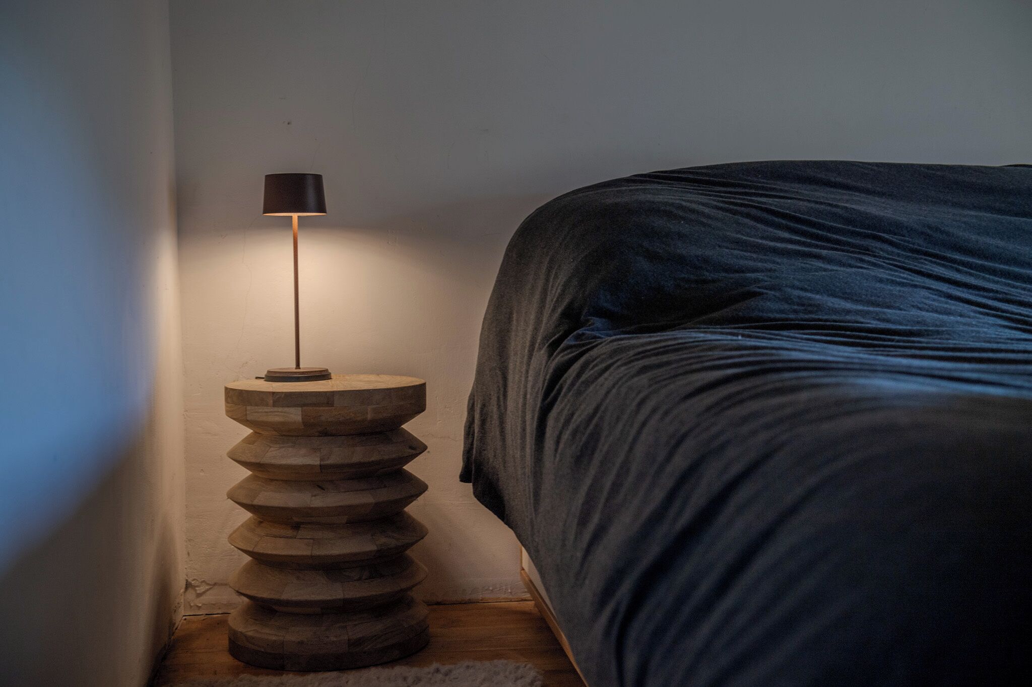 Un lit recouvert d’une couverture bleu foncé, une table de chevet ronde en bois avec une lampe noire, et un mur blanc.