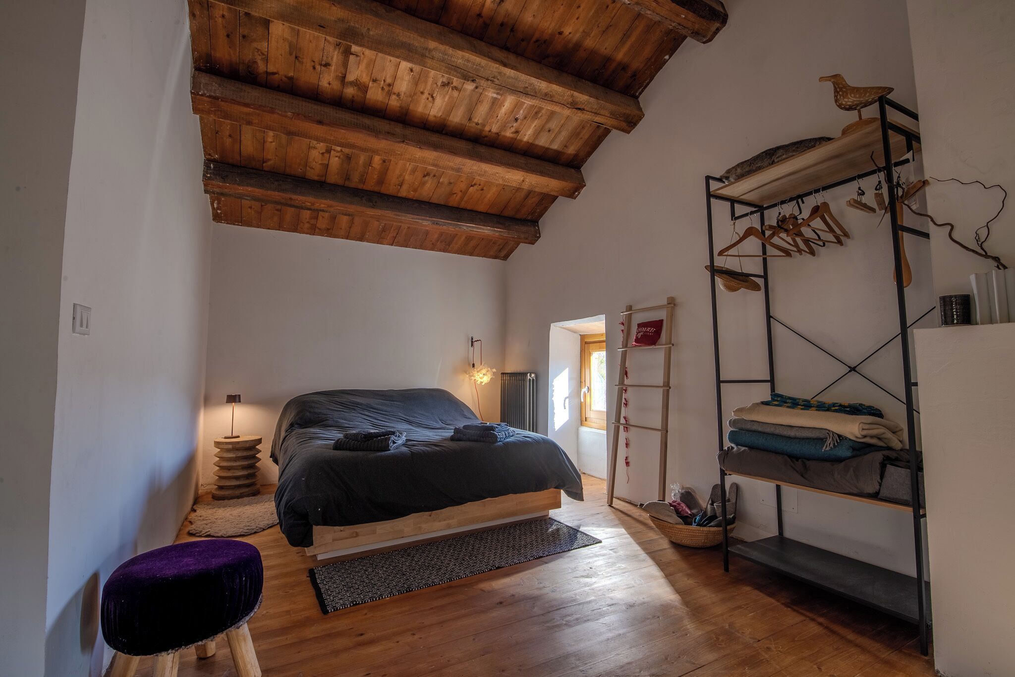 Une chambre avec un plafond en bois, un lit, un petit tabouret, une étagère à chaussures et un porte-objets fixé au mur pour y suspendre des vêtements.